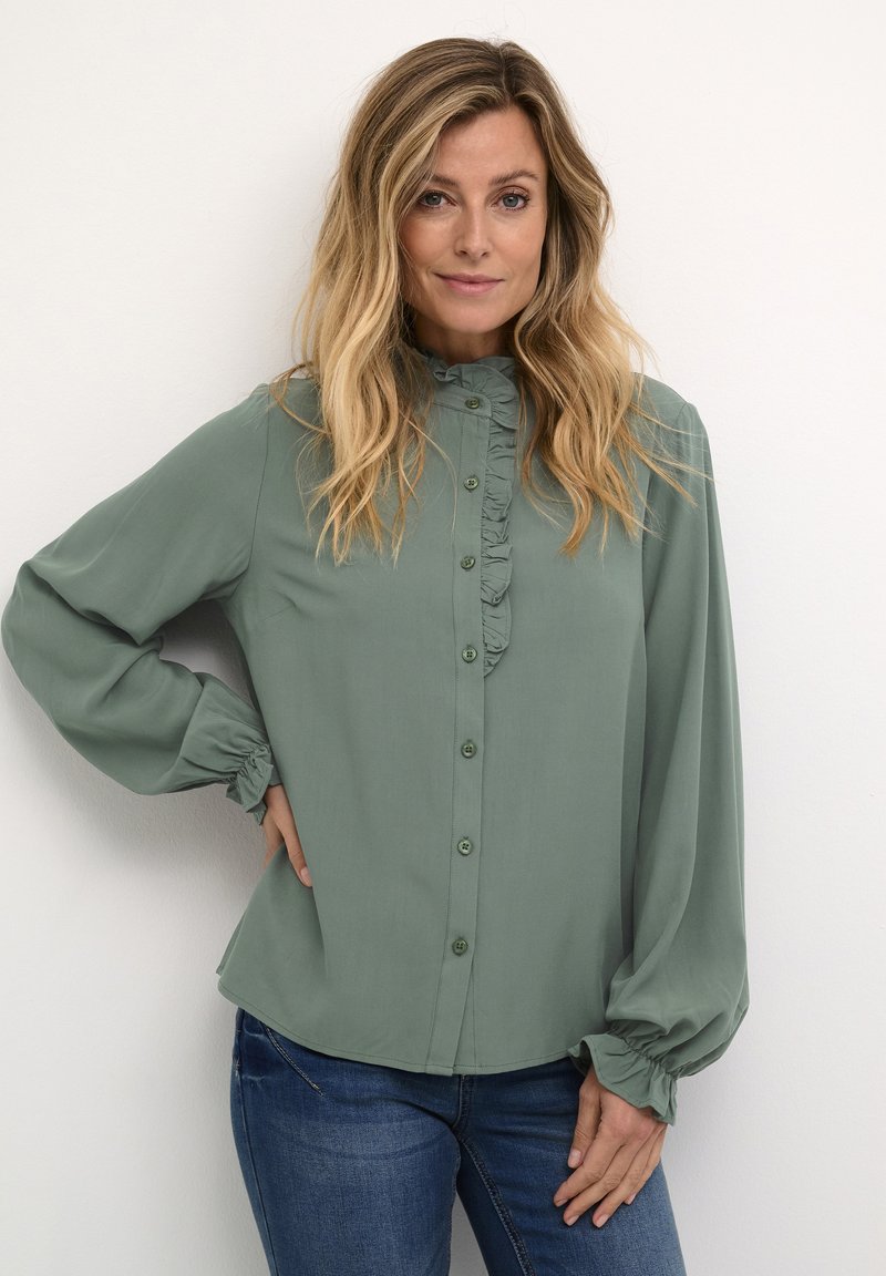 Blusa verde de manga larga con detalles de volantes a lo largo del placket de botones. Tejido suave, puños ajustados y un brillo sutil, combinada con jeans azules.