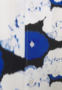 Tissu de chemise blanche avec des motifs abstraits de coups de pinceau bleus et noirs, présentant des boutons blancs le long de la couture centrale.