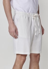 Uomo che indossa pantaloni corti bianchi con coulisse e tasche laterali, con una t-shirt bianca infilata, in piedi davanti a uno sfondo neutro.