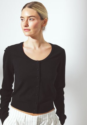 Zwarte cardigan met een ronde halslijn, knoopsluiting aan de voorkant, getailleerd ontwerp en lange mouwen, gecombineerd met witte streepjesbroek.