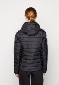 Svart pufferjacka med huva, med vita sidostripes, quiltad textur och elastiska ärmslut. Bakåtsyn visar den figursydde designen.