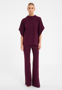 Bordeauxfarbener Zweiteiler bestehend aus einem locker sitzenden Oberteil mit hohem Kragen und weiten Ärmeln, kombiniert mit langen, eng anliegenden Hosen. Glatte Textur.