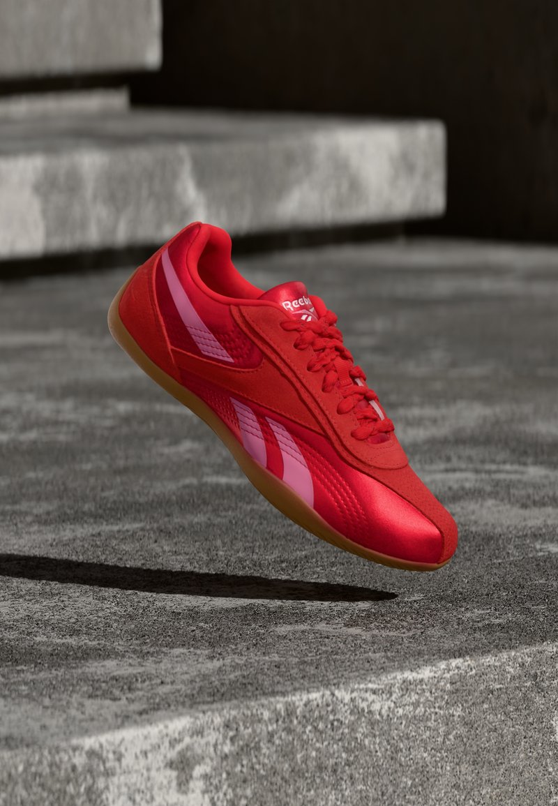 Roter Reebok-Sneaker mit pinken Streifen und Gummisohle, der über grauen Betontreppen schwebt, die im Schatten liegen.