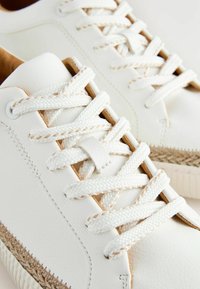 Sneaker bianchi in pelle texturizzata, con lacci crema, un accento marrone chiaro e dettagli intrecciati lungo la suola. Design complessivamente pulito.
