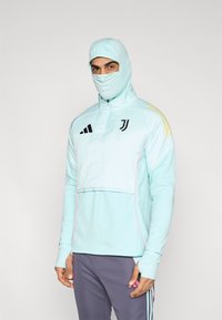 Lyseblå atletisk hættetrøje med høj krave og tætsiddende hætte, med sort Adidas-logo og gule accenter, båret over grå bukser.