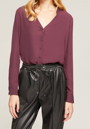 Mujer vistiendo una blusa morada de manga larga con botones forrados de tela y pantalones de cuero negro con cintura elástica y cordón ajustable.