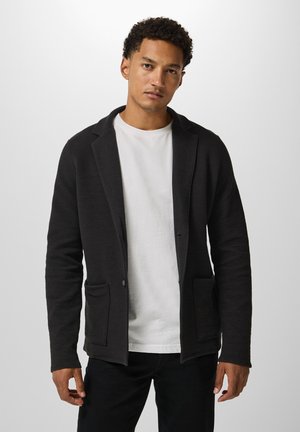 Schwarze Strickblazer mit weicher Textur, Reverskragen, zwei vorderen Taschen und einem einzelnen Knopfverschluss, über einem weißen T-Shirt getragen.