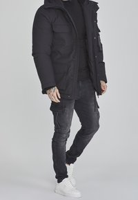 Musta toppatakki hupulla, jossa useita etutaskuja ja vetoketjusuljenta. Yhdistetty tummiin cargo-housuihin ja valkoisiin tennareihin.