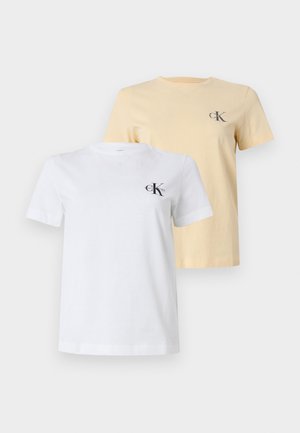 Γυναίκα που φοράει λευκό T-shirt με λογότυπο CK και μαύρο τζιν με φαρδιά πόδια, στέκεται δίπλα σε άσπρο τοίχο με γκρι δάπεδο.