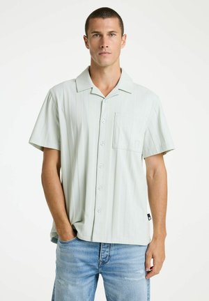 Muž s krátkymi vlasmi, oblečený v svetlom krátkom rukáve s gombíkmi a modrých džínsach, stojaci s jednou rukou v vrecku na bielom pozadí.