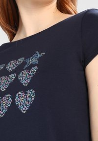 Marineblå skjorte med seks blomstermønstrede hjerter og en blomstermønstrede stjerne nær halsen, båret af en person med lys hud.