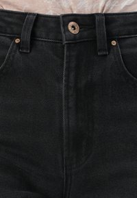 Primer plano de la cintura de unos pantalones de mezclilla negros con un botón, presillas para el cinturón y bolsillos visibles sobre un fondo neutro.