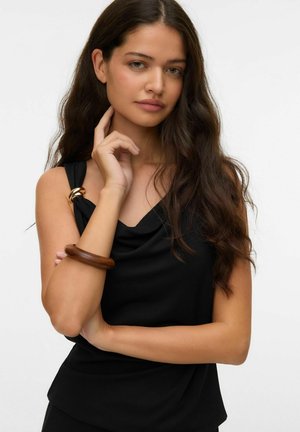 Giovane donna con lunghi capelli castani che indossa un top nero senza maniche con dettaglio ad anello dorato e un bracciale di legno, posando con la mano vicino al viso.