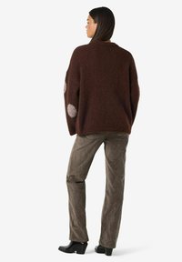 Kvinna som står, iklädd en mörkbrun oversized tröja med ljusgrå armbågsveck, grå kordflodbyxor och svarta klackstövlar.