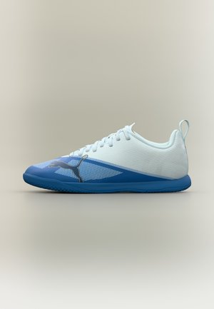 Chaussure de football en salle Puma blanche et bleue, avec des lacets, une tige texturée et une semelle en caoutchouc bleue, présentée de profil sur un fond uni.
