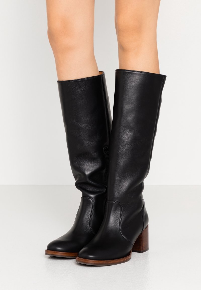 Bottes hautes noires en cuir avec un bout arrondi, un talon bloc et une texture lisse. Fermeture éclair latérale. Détail en bois naturel sur le talon.