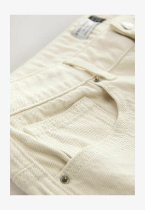 Kremo spalvos medvilniniai kelnės, turinčios tekstūruotą audinį, matomas kišenes ir metalinį mygtuko akcentą su etikete, matoma palei juosmenį.