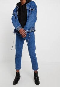 Person stands wearing blue denim jacket and pants, black hoodie, and black ankle boots with buckles on plain white background.