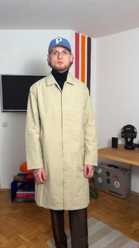 Jauns vīrietis, valkājot bēšas krāsas garu mēteli, melnu augstu apkakli, zilu cepuri, stāv istabā ar krāsainām svītrām, televizoru, galdu ar austiņām un koferi.