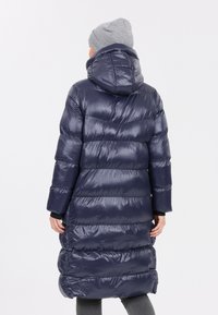 Marinblå pufferjacka med huva, som har en blank, quiltad textur, elastiska ärmslut och en lång silhuett, ihop med en grå mössa.