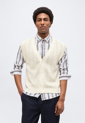 Jeune homme aux cheveux bouclés portant un gilet beige en tricot par-dessus une chemise rayée à boutons avec les manches retroussées et un pantalon foncé.