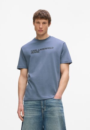 Jeune homme portant un t-shirt bleu Karl Lagerfeld Jeans et un jean en denim bleu délavé, debout devant un fond clair uni.