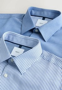 Dve lepo zloženi moški srajci, ena svetlo modra enobarvna in ena bela z mornarsko modrimi črtami, obe z ovratniki na gumbe in oznakami Next.
