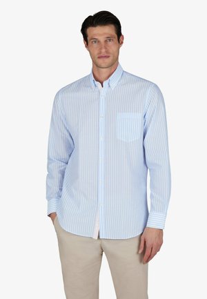 Homme portant une chemise rayée bleu clair et blanc avec poche poitrine et pantalon beige, debout avec une main dans la poche.