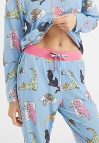 Niebieski zestaw piżamowy z zabawnym wzorem kotów. Górna część ma długie rękawy; spodnie mają różową talię z sznurkiem do regulacji oraz elastyczny krój.