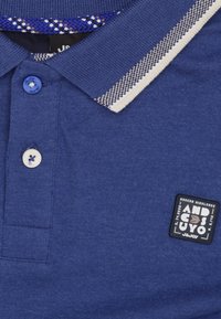 Blauwe polo met een gestreepte kraag, twee knopen en een geweven patroon aan de binnenkant. Een genaaid etiket is geplaatst boven de onderzoom.