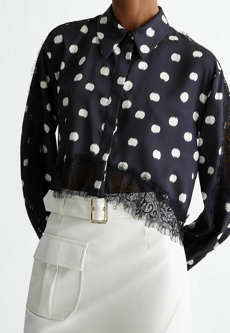 Blouse noire boutonnée avec des pois blancs et un bord en dentelle, associée à une jupe blanche taille haute avec une grande poche et une ceinture.
