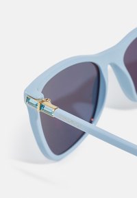 Ochelari de soare de culoare albastru deschis, cu lentile întunecate, având accesorii din aur și accente clare de cristale pe temple. "SWAROVSKI" gravat pe lateral.