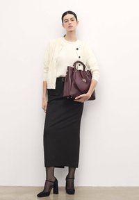 Structured handbag in dark burgundy leather, featuring gold hardware. Woman in cream cardigan and black skirt, holding the bag.