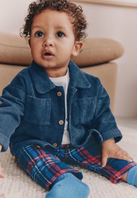 Chaqueta azul de pana con botones y dos bolsillos delanteros, combinada con pantalones a cuadros rojos y azules. El niño tiene el pelo rizado y lleva una camisa blanca.