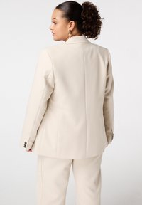 Vrouw met krullend haar in een paardenstaart, gekleed in een crèmekleurige op maat gemaakte blazer en bijpassende broek, iets zijwaarts gekeerd tegen een witte achtergrond.