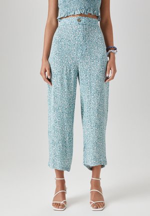 Mujer con pantalones florales de cintura alta en azul claro recortados por encima de los tobillos, combinados con sandalias blancas con tiras y una camiseta azul a juego.