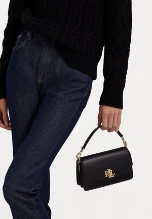 Person iført mørkeblå jeans og sort kabelstrikket sweater, der holder en lille sort læderhåndtaske med gyldt RL-logo.