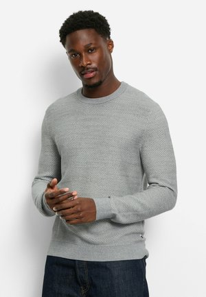 Light gray knitted crewneck sweater with textured pattern, ribbed cuffs, and hem; worn over dark jeans, hands positioned at front.
