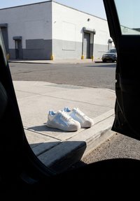 Hvide og blå sneakers placeret på en fortovskant set indefra en åben bildør på et solrigt gadehjørne.