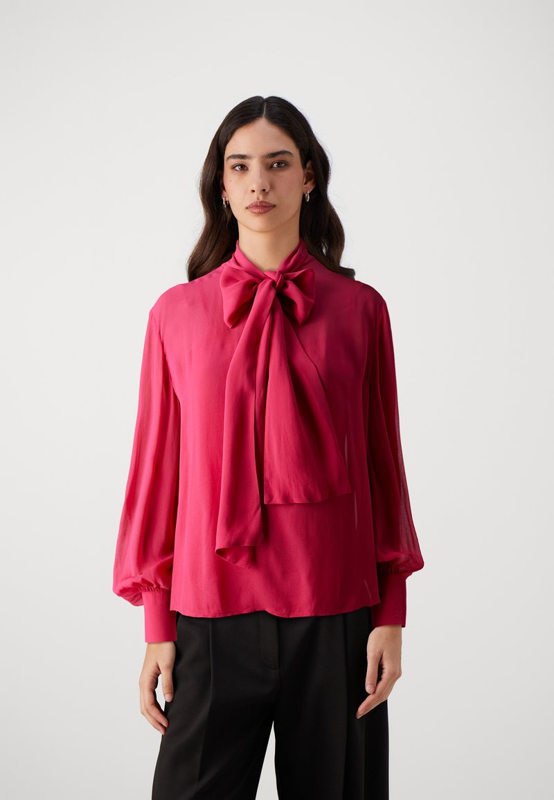 Fuchsia silkebluse med stort sløjfe-krave, lange pufærmer og en afslappet pasform. Sammen med sorte bukser. Glat tekstur.