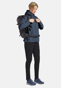 Giacca impermeabile navy, zaino nero con cinture, pantaloni neri e scarpe atletiche blu. Texture liscia, design ergonomico, dettagli funzionali.
