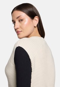 Pull en maille crème avec un motif texturé, présentant une couche noire à manches longues en dessous. Le matériau semble doux et respirant.