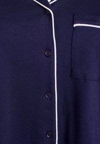 Camicia blu navy con bottoni, dettagli in piping bianchi, dotata di taschino sul petto e bottoni rotondi viola, realizzata in tessuto morbido.