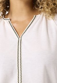 Biała bluzka z dekoltem w kształcie litery V, ozdobiona czarnym i złotym haftem dekoracyjnym wzdłuż listwy guzikowej. Lekka tkanina, gładka tekstura.