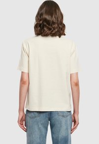Personne avec des cheveux bruns ondulés mi-longs portant un t-shirt à manches courtes beige uni et un jean en denim bleu, vue de dos.