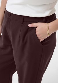 Burgundy tailored pants with a smooth texture, featuring a button closure, side pockets, and an elastic waistband detail.