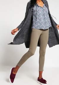 Cardigan long gris, blouse fleurie bleue, pantalon beige ajusté avec des fentes aux chevilles, et chaussures slip-on bordeaux avec un détail noué.