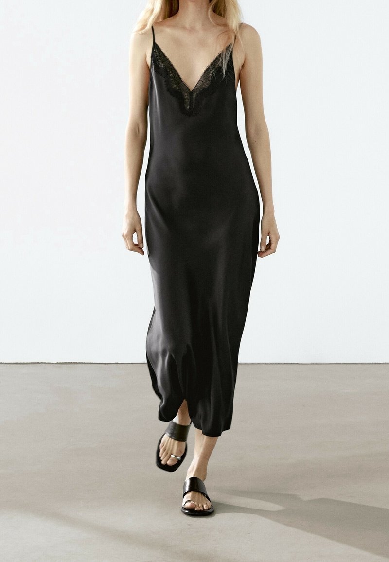 Femme marchant portant une robe noire sans manches avec bordure en dentelle et des sandales noires à bout ouvert sur un sol lisse avec un fond blanc uni.