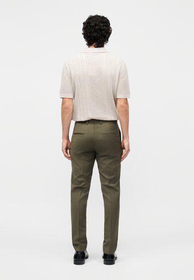 Άνδρας με σγουρά μαλλιά που φοράει μπεζ πλεκτό με κοντά μανίκια, ελιάς παντελόνια και μαύρα παπούτσια, στέκεται γυρισμένος με την πλάτη στο μονόχρωμο φόντο.