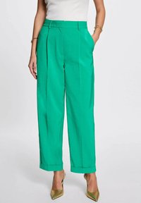 Pantaloni plissettati verdi con vestibilità sartoriale, orlo con risvolto e texture liscia, abbinati a tacchi a punta dorati.